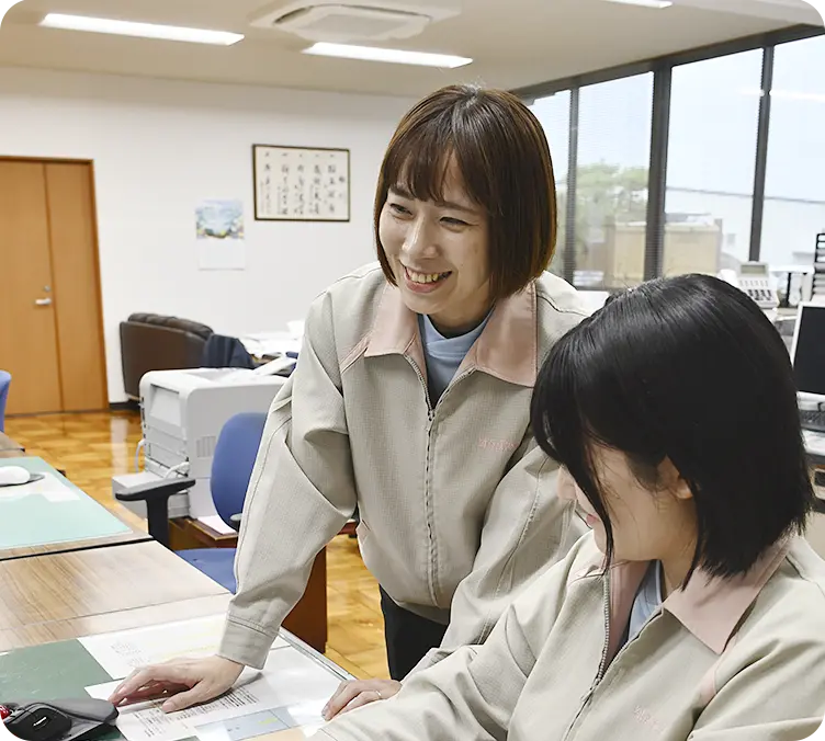 オフィスで女性社員2人がパソコン画面を見ながら打ち合わせをしている様子。先輩社員が笑顔で後輩にアドバイスしている。