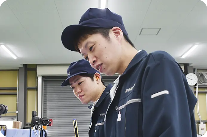 作業着姿の男性社員2人が機械の前で真剣に作業内容を確認している。工場内での製造・検査の様子。